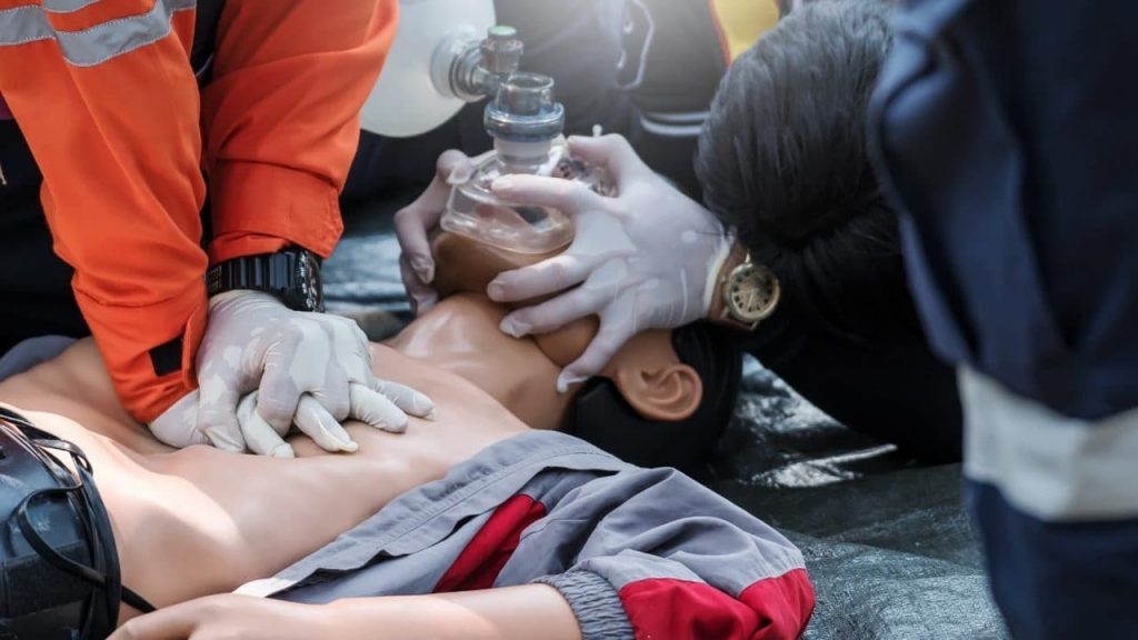 A person wearing an orange jacket performs CPR on a mannequin, compressing the chest, while another individual in dark clothing holds an oxygen mask over the mannequin's face during a training session. Both wear protective gloves.