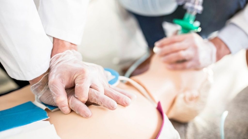 A person wearing gloves is performing chest compressions on a dummy during a CPR training session while another individual practices giving breaths using a resuscitation mask. The image focuses on the hands on the dummy's chest and the mask on its face.