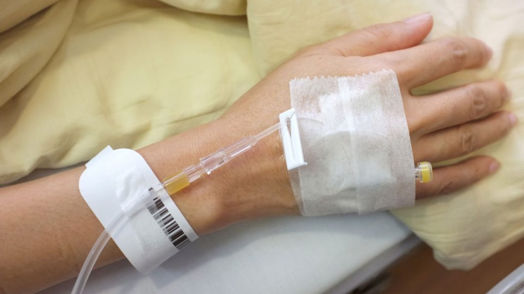 A close-up of a person's hand with a medical IV line inserted into the back of it, secured with white adhesive tape. The person is resting on a beige blanket, and a hospital wristband is visible around their wrist.