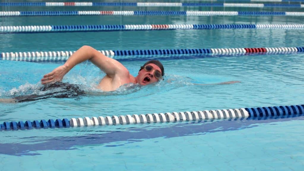 A swimmer wearing goggles and a swim cap performs a freestyle stroke in a swimming pool. The pool is divided into lanes with blue, white, and red lane dividers. The water is clear, and the swimmer is in the middle of a stroke with one arm extended forward.