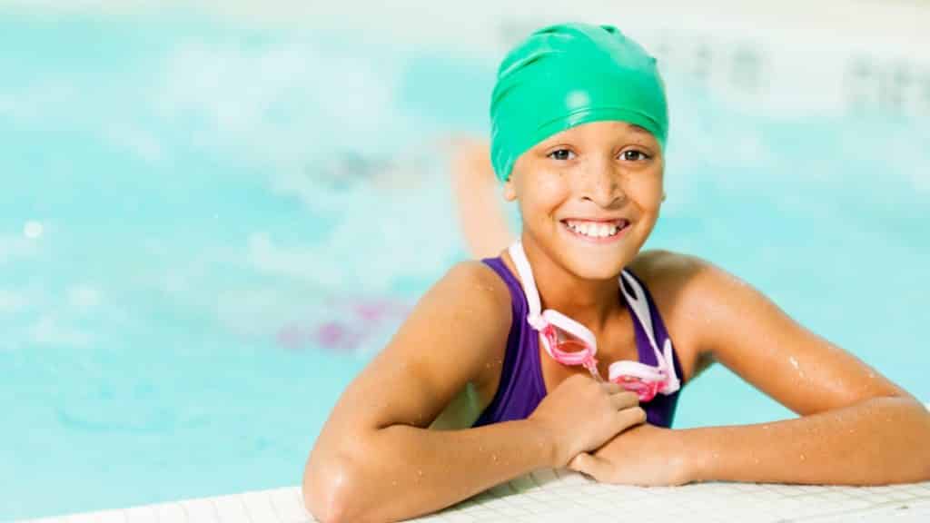 A smiling child wearing a green swim cap and pink goggles rests on the edge of a swimming pool. The water is a bright blue, and the child appears to be in a swimsuit, enjoying their swimming lesson in the pool.