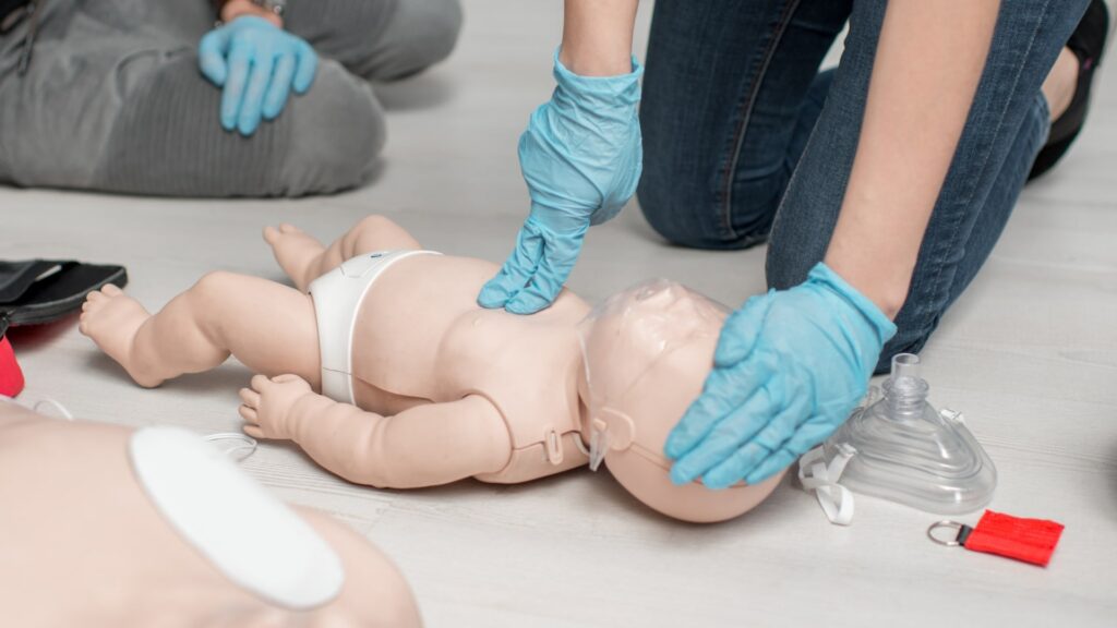 Two individuals wearing blue gloves practice CPR on an infant mannequin. One person performs chest compressions, while the other prepares a resuscitation mask next to the mannequin. Another adult mannequin torso is partially visible.