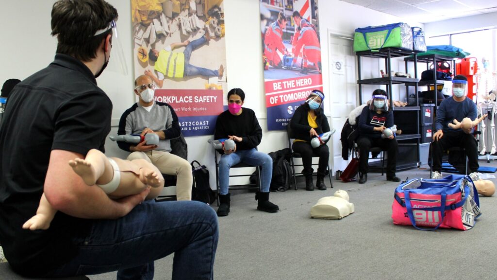 A group of people wearing masks and seated in a classroom are being instructed in infant CPR by a person holding a baby mannequin. Various training equipment and instructional posters are visible in the background.