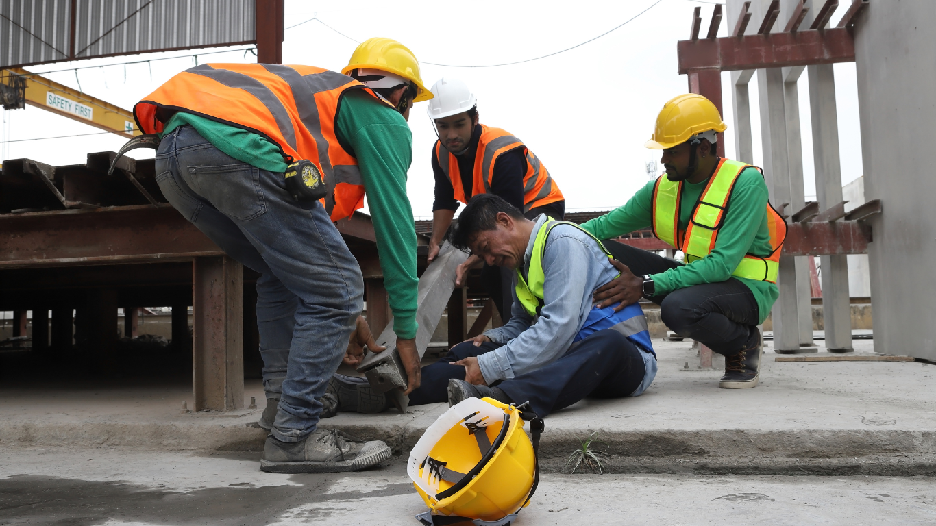 A group of construction workers in safety vests and helmets attending to an injured colleague on a construction site, with a 'SAFETY FIRST' banner in the background
