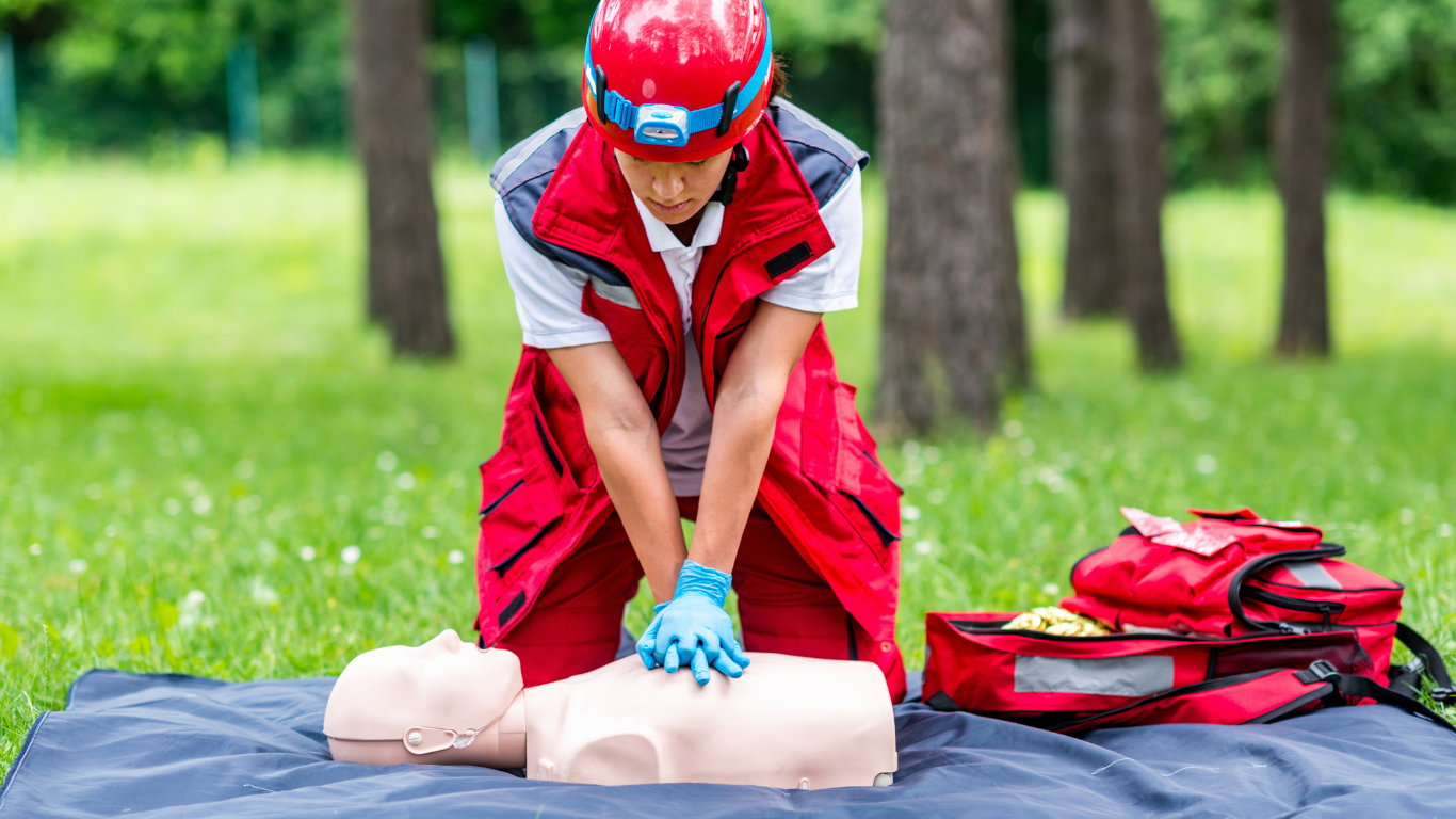 A first aid instructor in a red vest and helmet performing CPR on a training manikin in a grassy outdoor area, with a medical kit open nearby