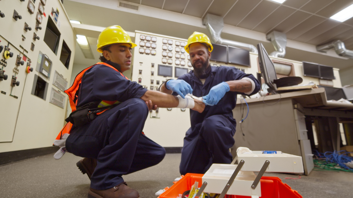 Two construction workers in a control room, with one worker administering first aid by bandaging the arm of the other worker, both wearing hard hats and safety gear