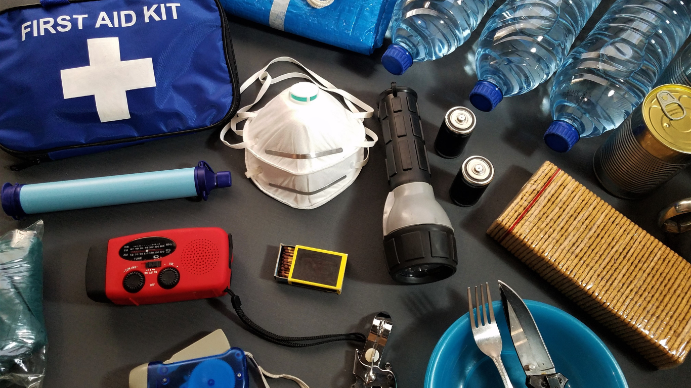 A first aid kit and various emergency supplies, including a flashlight, water bottles, face masks, batteries, and a portable radio, laid out on a flat surface