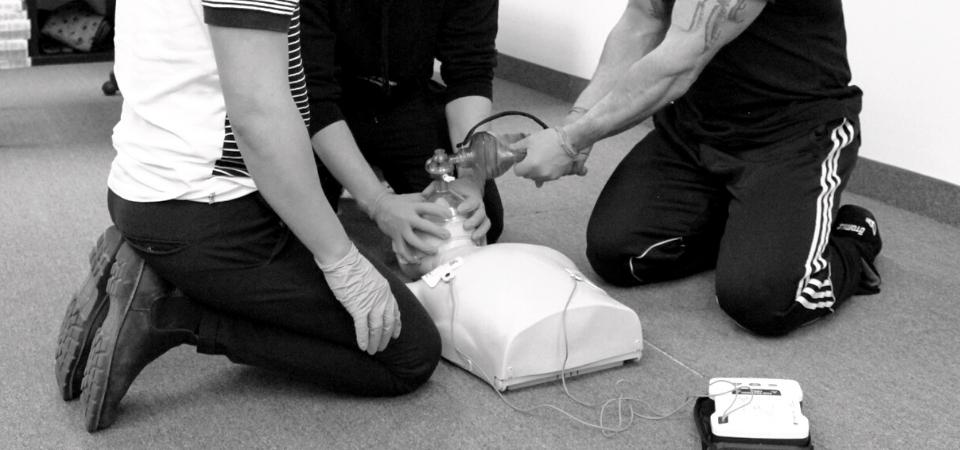 Three individuals are kneeling on the floor, practicing CPR on a mannequin. One person is using a bag valve mask, another assists in holding the mask, and the third monitors an AED device. They are all focused on the resuscitation exercise.