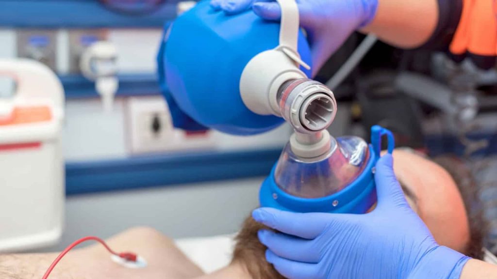 A medical professional wearing blue gloves administers ventilation to a patient using a manual resuscitator in a clinical setting. The patient's face is partially covered by the resuscitator mask, with medical equipment visible in the background.