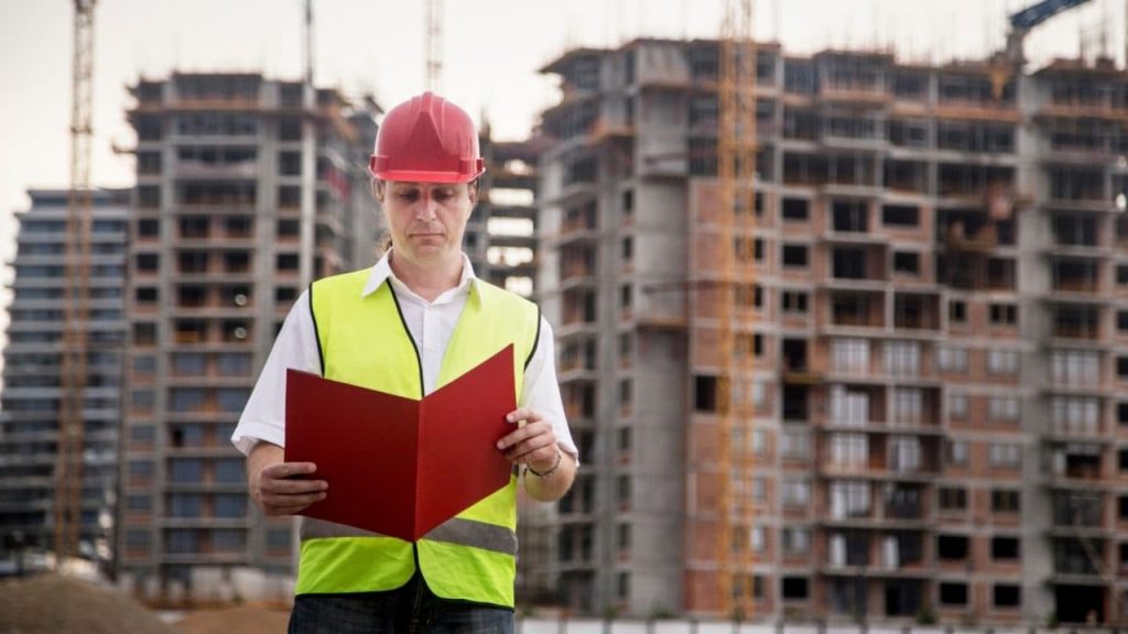 A construction worker in a red hard hat and yellow safety vest stands in front of tall, unfinished buildings, holding and reading a red folder. Cranes are visible in the background under a clear sky.