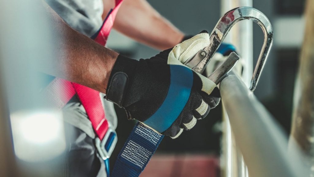 A person wearing gloves is securing a safety harness to a metal railing. The image focuses on their hands holding a hook, emphasizing construction safety equipment.