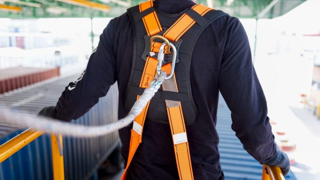 A person wearing an orange and black safety harness is attached to a secure line. They are standing on an elevated platform with blue shipping containers visible below.