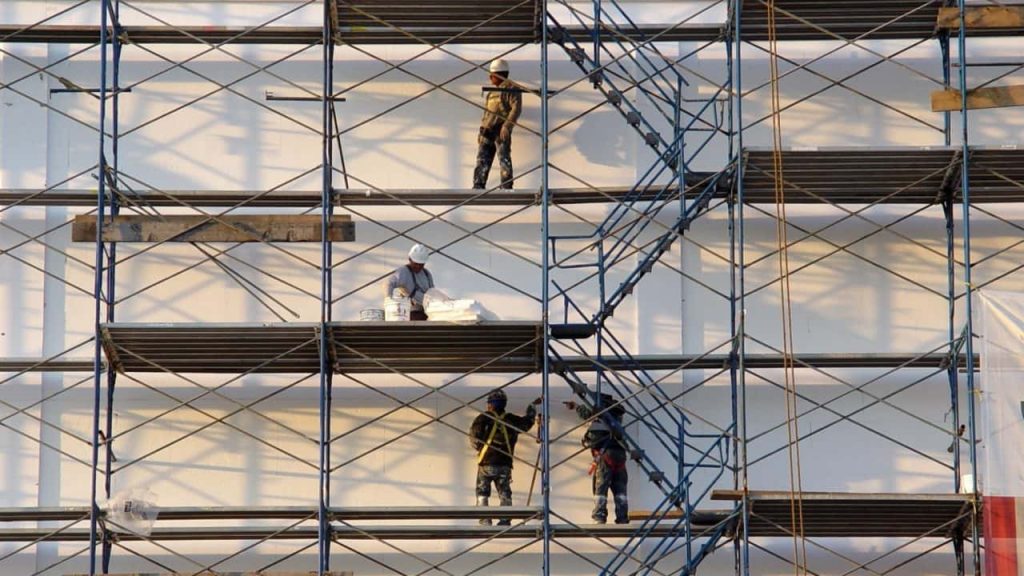 Construction workers in safety gear stand on a large metal scaffold against a white building. One worker is carrying materials, while others are stationed on different levels of the scaffolding.