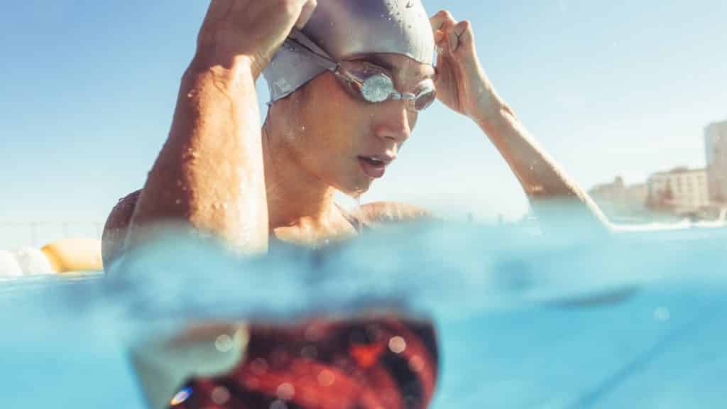 A swimmer wearing a swim cap and goggles adjusts her gear while standing in a pool. The water level is midway up her body, and she appears to be focused. The background shows a clear blue sky and some buildings in the distance.