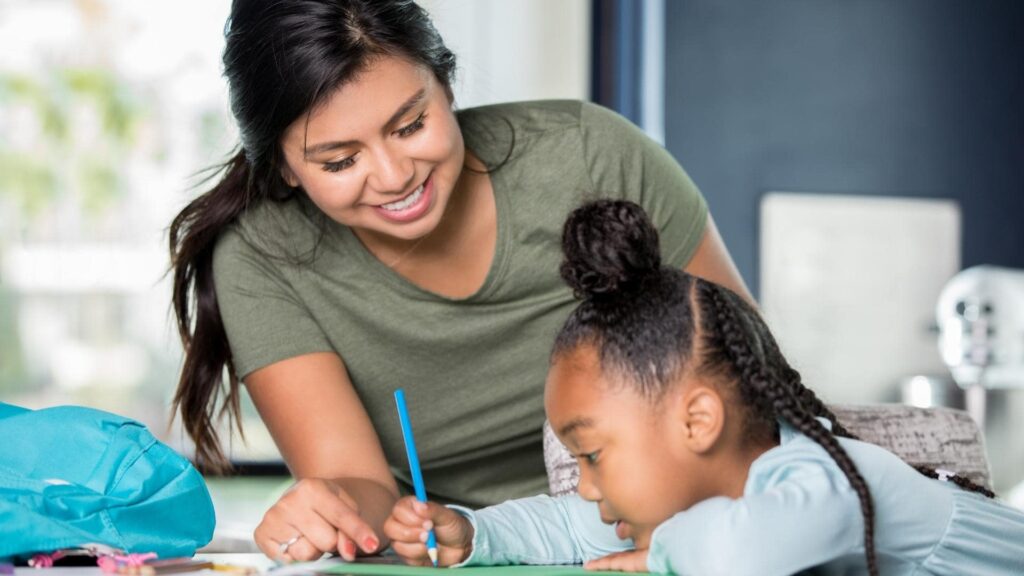 A woman in a green shirt smiles while helping a child with braided hair and a top bun, who is writing with a pencil. They are sitting at a table with papers and a blue bag nearby. The setting appears to be an indoor educational environment.