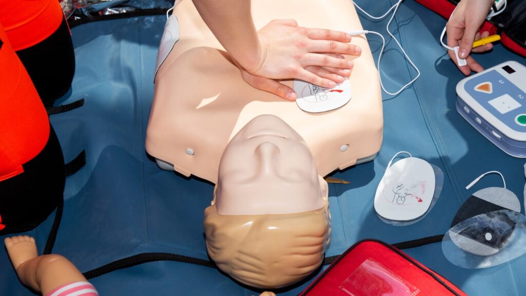 A person practices CPR on a medical training dummy. The person's hands are placed on the chest of the dummy, which is equipped with defibrillator pads. Various CPR and first aid equipment are visible around the scene.