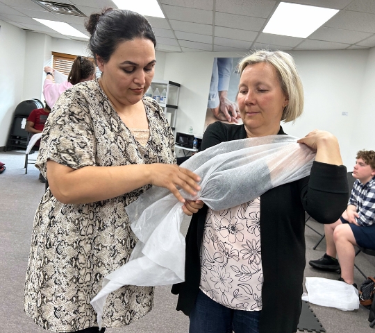 Two women in a room, one is adjusting a sheer white fabric on the other's arms. there are people and chairs in the background, suggesting a casual gathering or workshop setting.