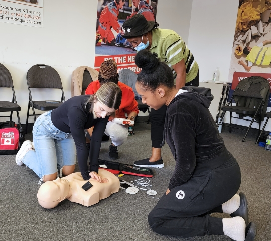 Three people practicing cpr on a dummy in a classroom setting, focusing intently on the task, with posters and chairs visible in the background.