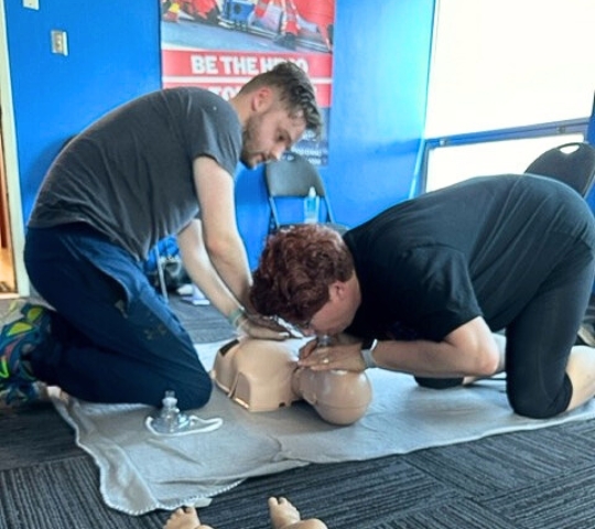 Two individuals practicing cpr on a mannequin in a room with blue walls and a 'be the hero' poster in the background.