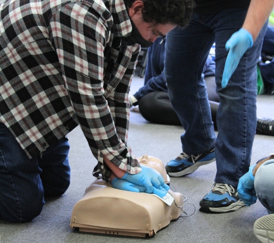 Two individuals practice cpr on a training mannequin on the floor, one compressing the chest and the other watching, both wearing gloves.