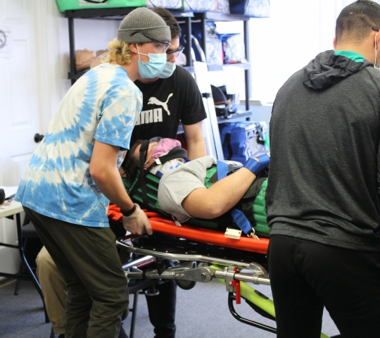 Three people in a room with medical equipment, one lying on a stretcher being attended to by two others, one in a tie-dye shirt and a mask.