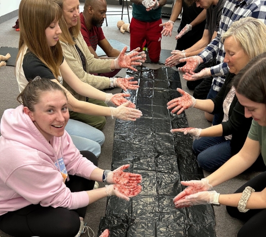 A group of diverse adults sits in a circle, smiling and interacting while pressing their red-painted hands onto a long black banner laid on the floor.