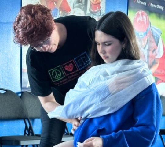 A woman in a blue top is being draped with a white blanket by another person with curly hair, in what appears to be a first aid training session.