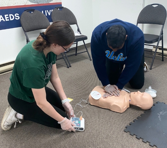 Two individuals, one in a green shirt and one in a blue ucla shirt, practicing cpr on a dummy in a classroom setting, using an aed training device.