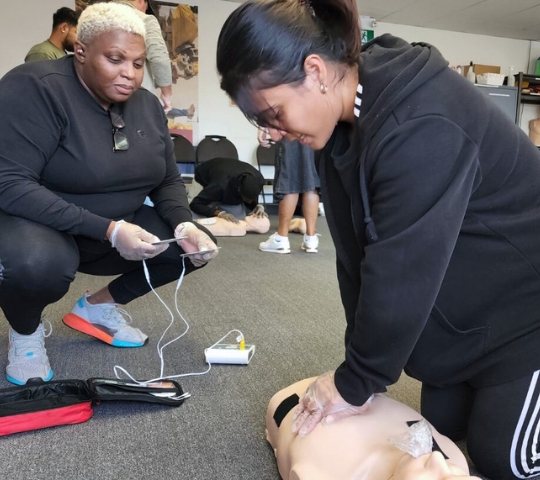 Two women practicing cpr on a mannequin in a training session, one holding defibrillator pads, in a classroom setting.