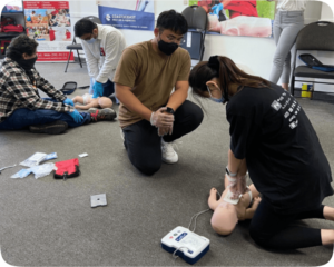 A CPR and first aid training session in progress with a man and a woman practicing on a dummy, while others observe and an instructor oversees. The room is equipped with life-saving materials.