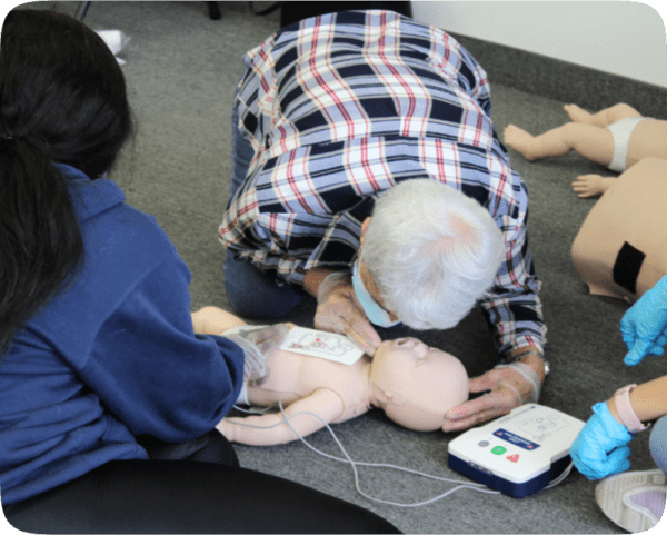 Elderly man and another individual practicing CPR on a dummy in a training session in Toronto, with a defibrillator and other dummies visible.