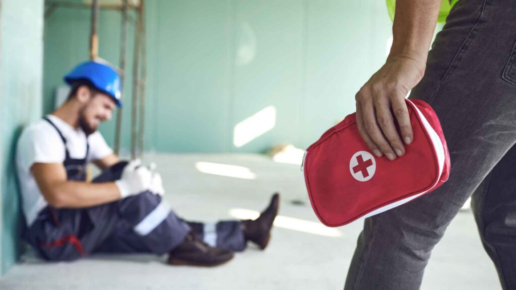 A construction worker sitting on the ground in the background with an injured leg, while another person approaches holding a red first aid kit