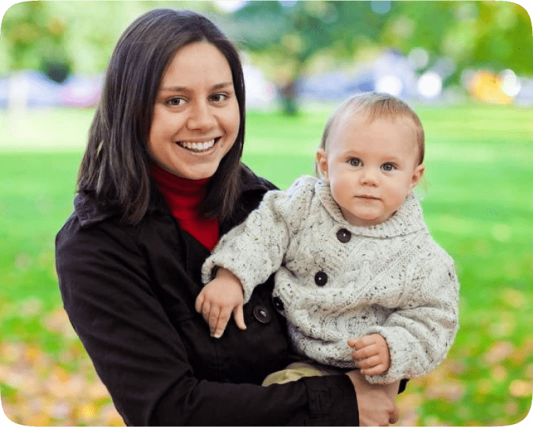 A smiling woman holds a baby in a knitted sweater, both looking at the camera with a blurred green park background.